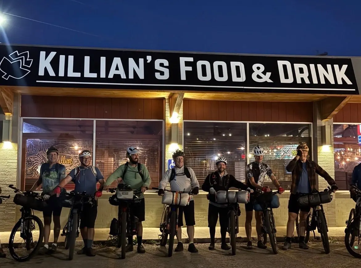 Seven cyclists with bikes outside the illuminated Killian's Food & Drink storefront at night in downtown Spearfish